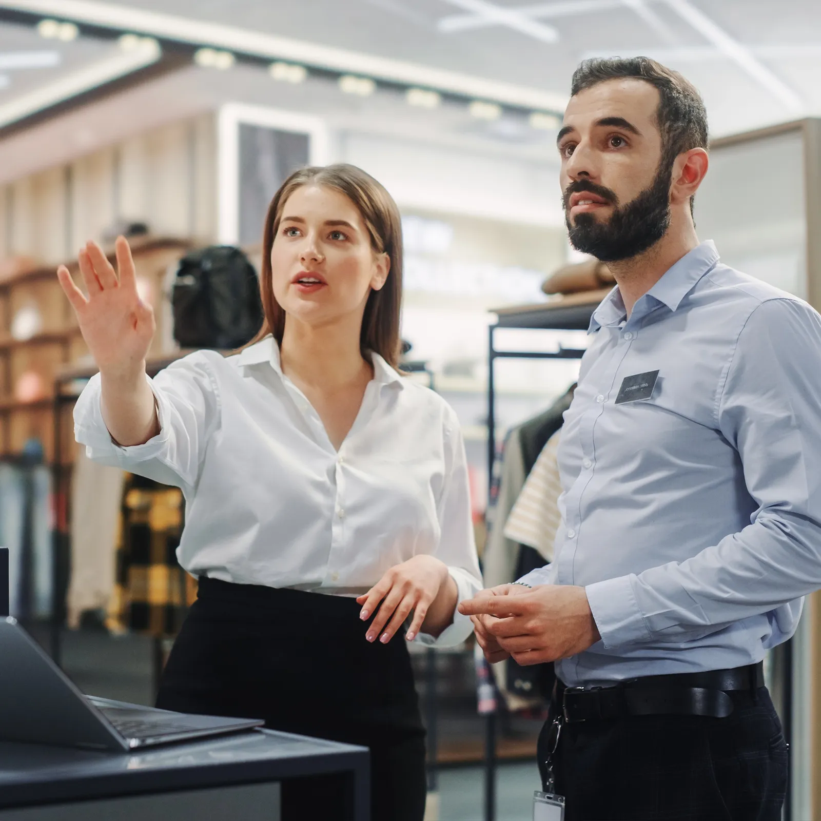 Two retail employees discussing near a laptop in a clothing store with racks in background.