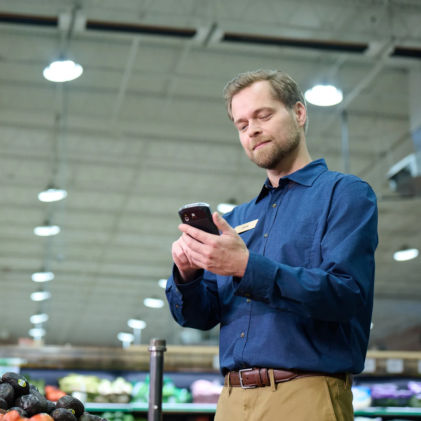 Man in blue shirt using a handheld device in a grocery store produce section.