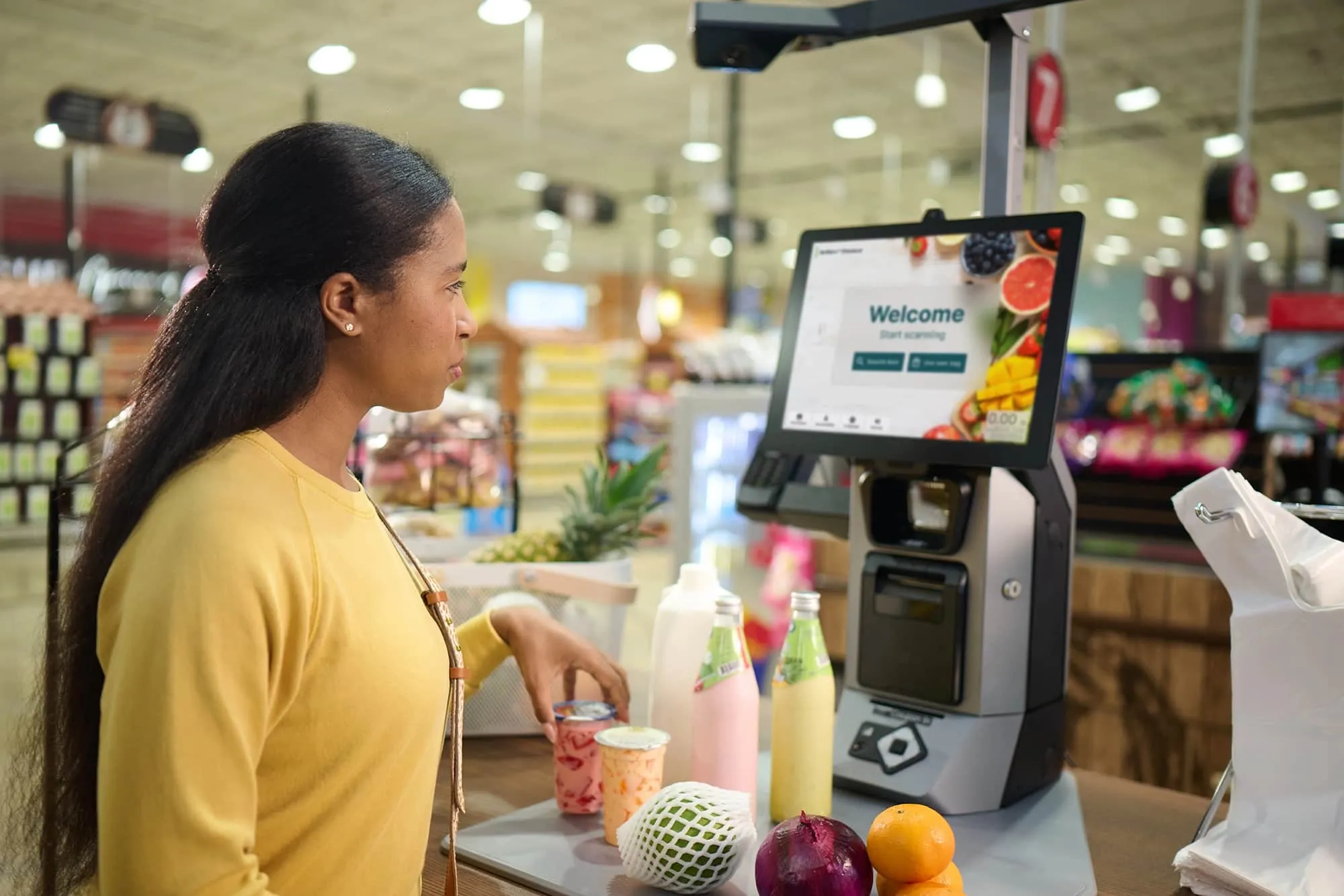 Woman using self-checkout machine with groceries including fruit, drinks, and an onion in a store.