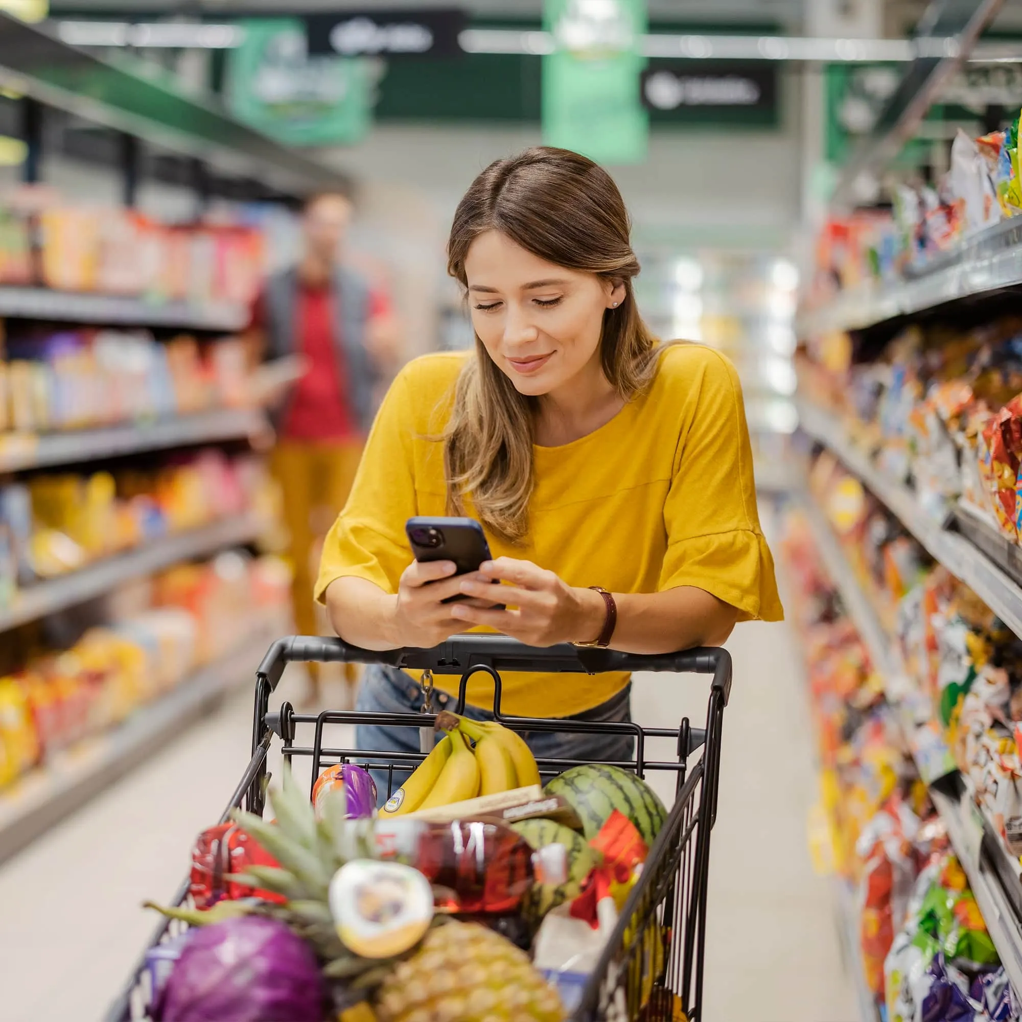 Woman in yellow shirt looking at phone while pushing a grocery cart filled with fruits in a supermarket aisle.