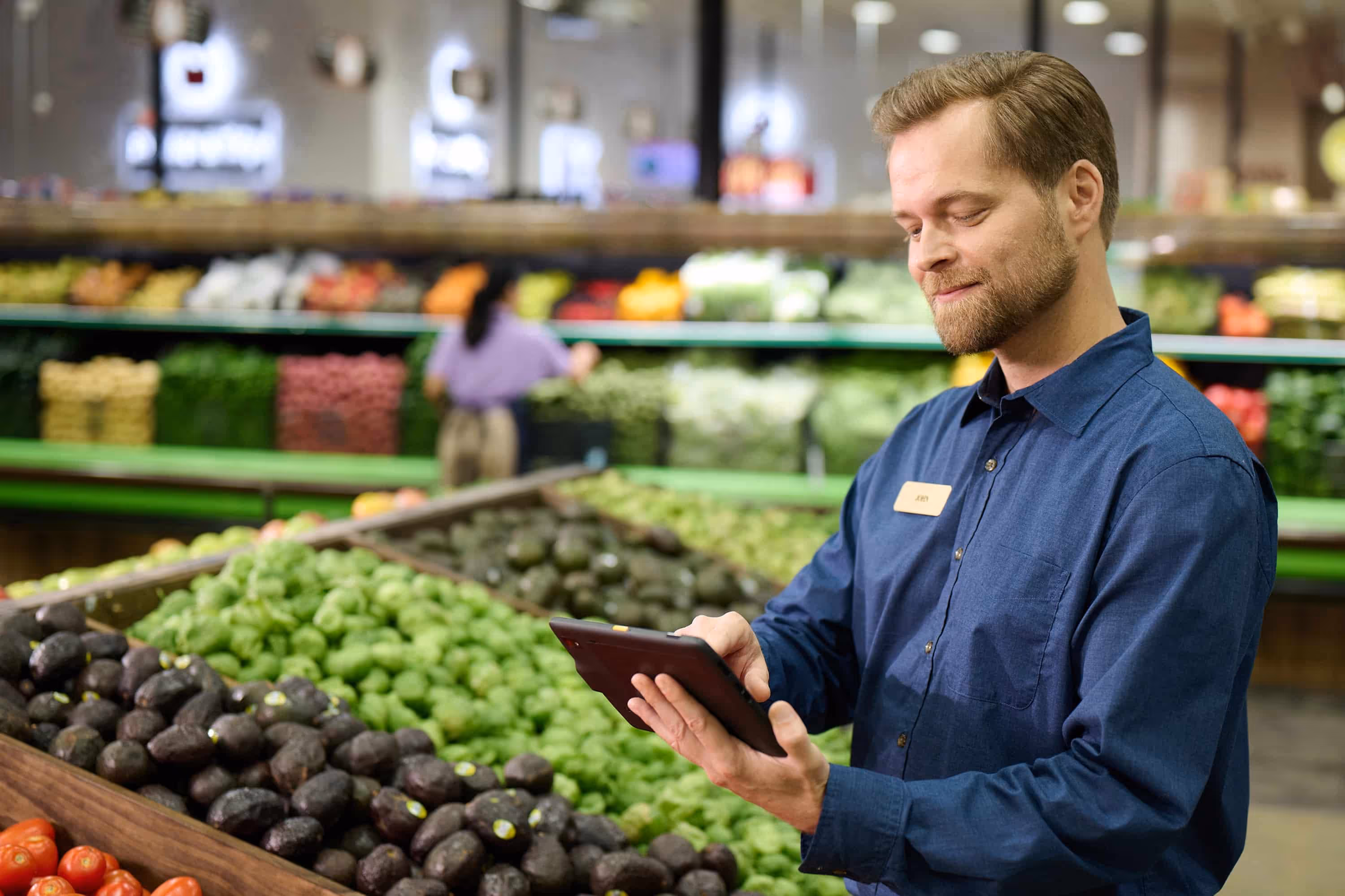 Man in blue shirt holding a tablet while standing by avocados and green produce in a grocery store.