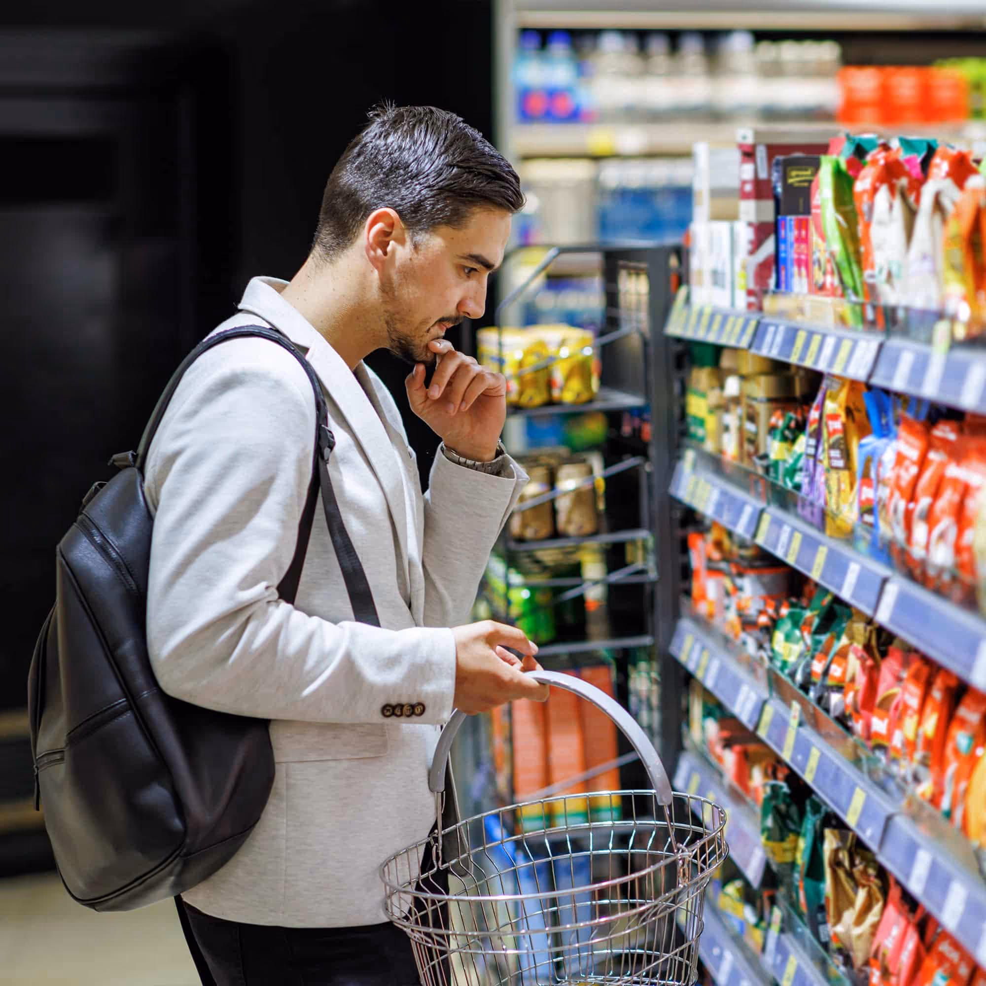 Man in light jacket with backpack holding empty shopping basket while browsing snacks in store aisle.