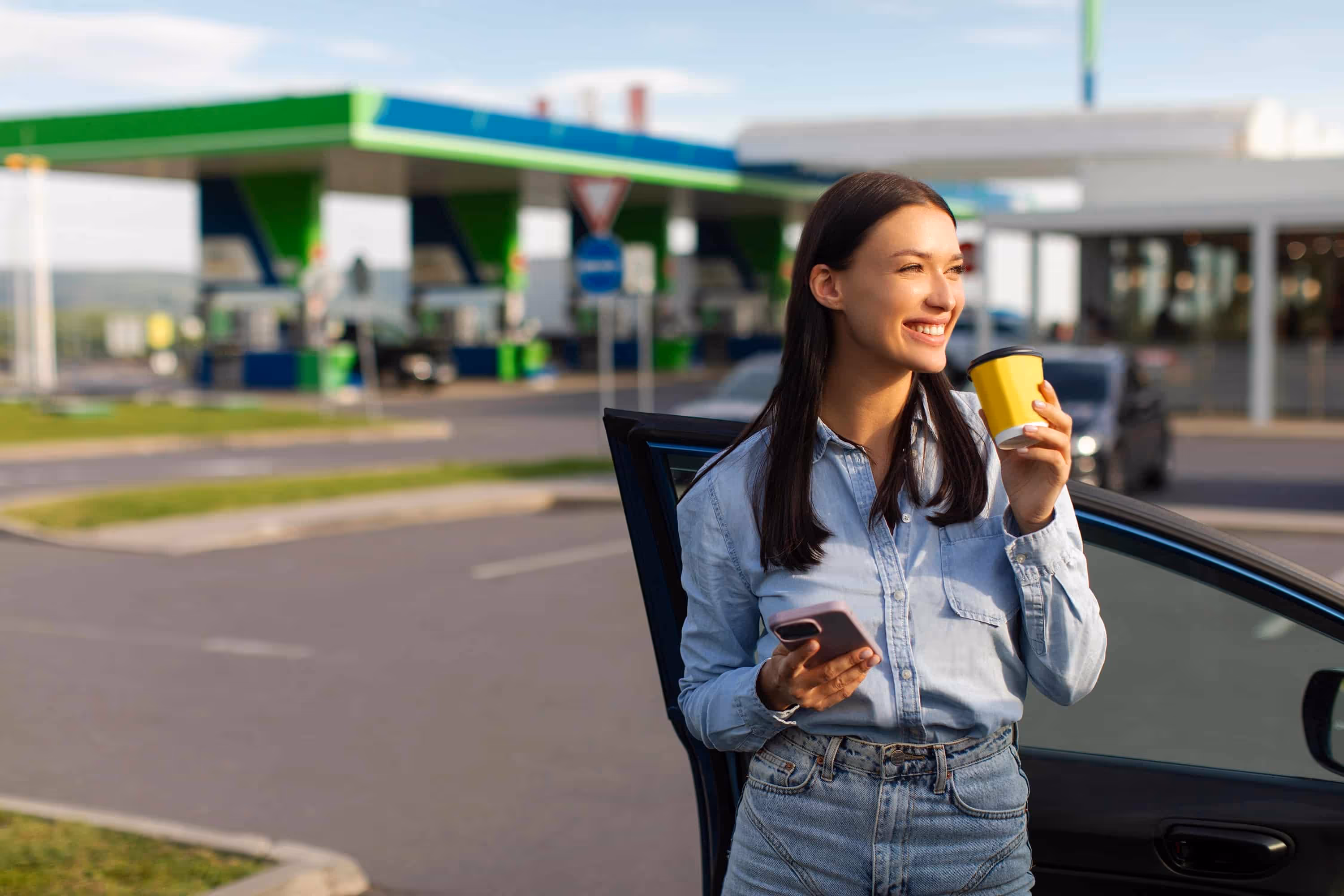 Smiling woman holding a yellow coffee cup and phone beside her car at a gas station.
