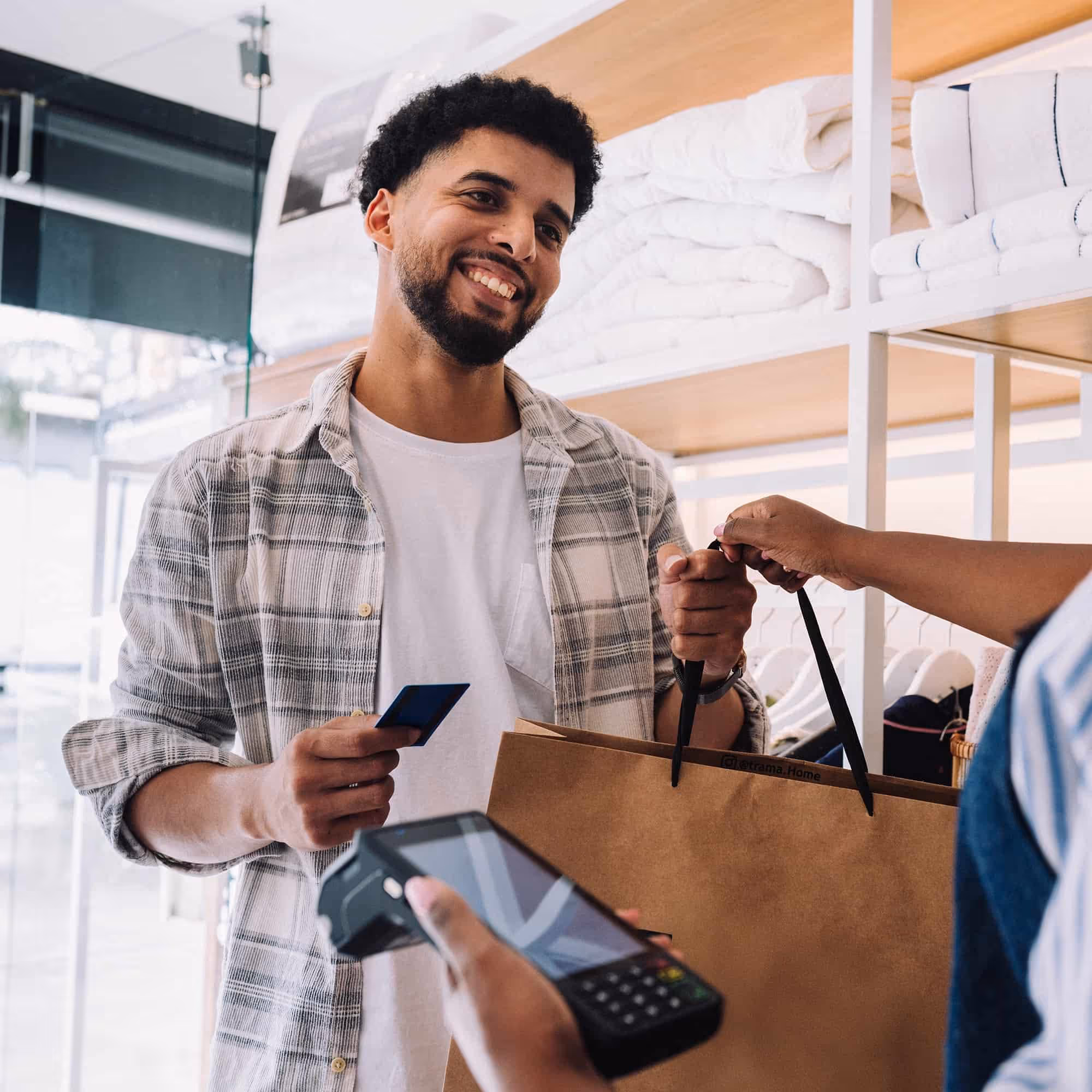 Smiling man holds credit card and shopping bag while cashier processes payment with card reader.