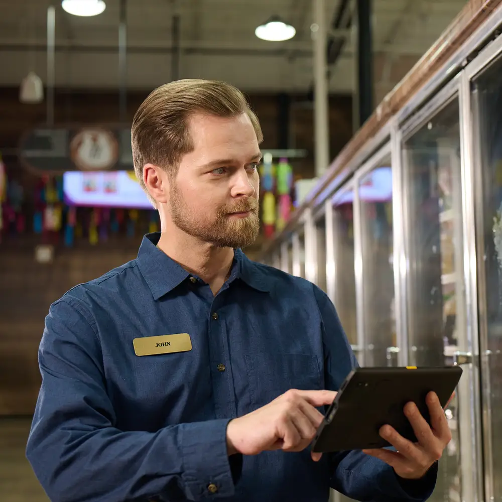 Man in a blue shirt and name tag using a tablet near freezer doors in a store.