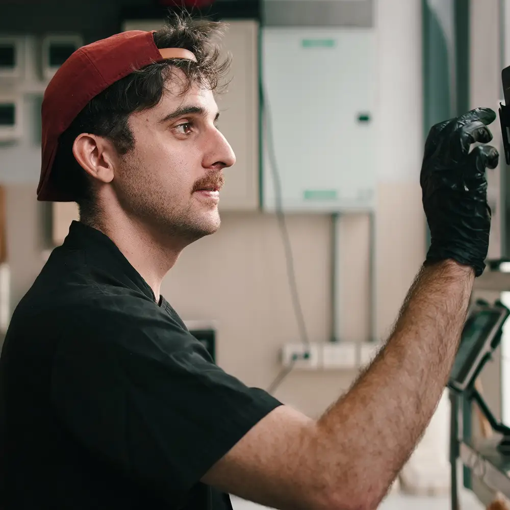 Man wearing a red backward cap and black gloves working on a machine or screen indoors.