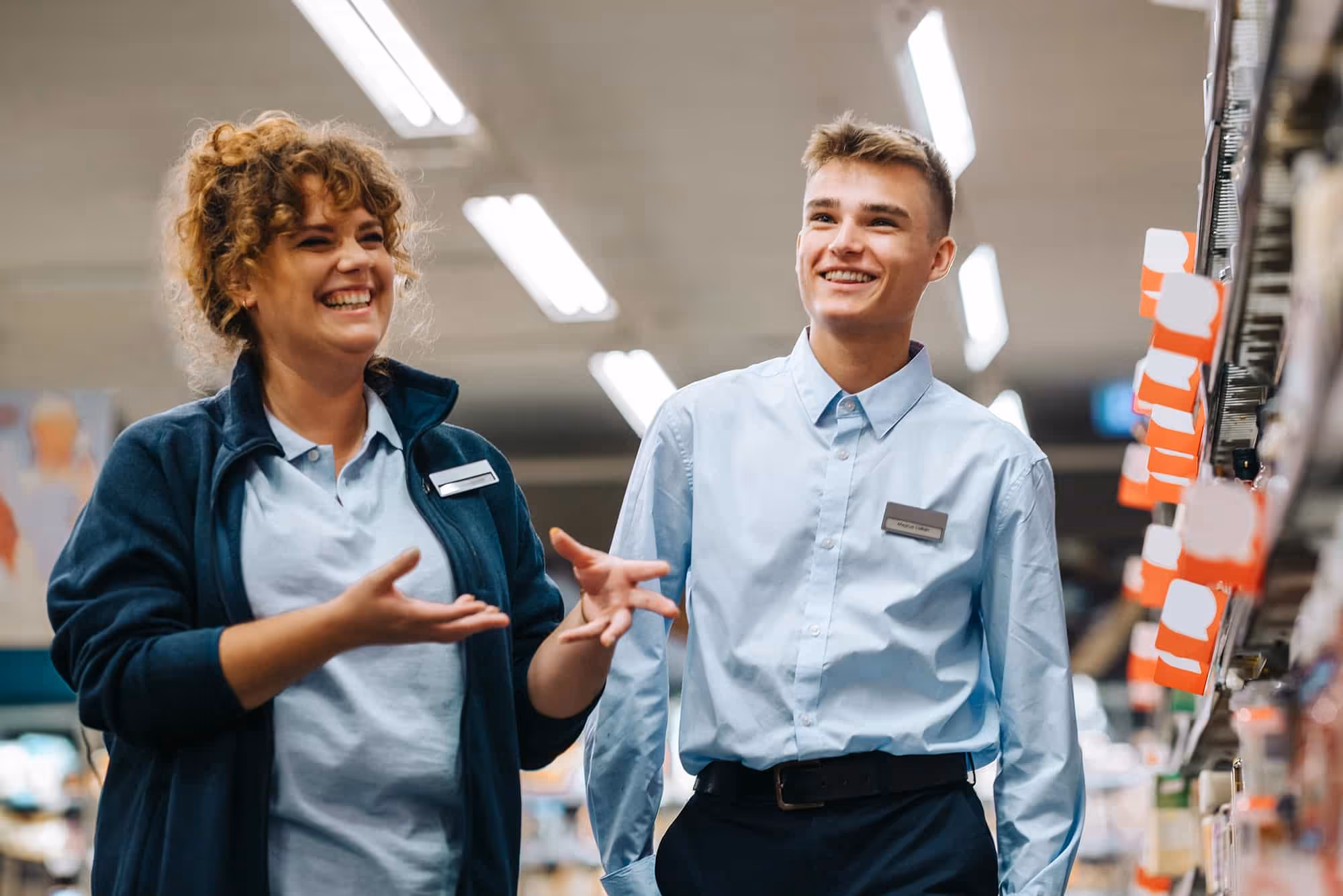 Two smiling store employees in uniforms standing and talking in an aisle with shelves and price tags.