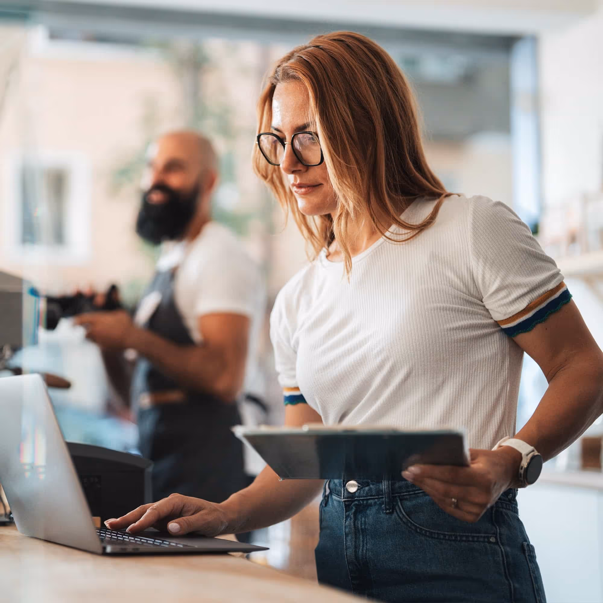 Woman with glasses using a laptop and holding a tablet in a cafe with a barista in the background.