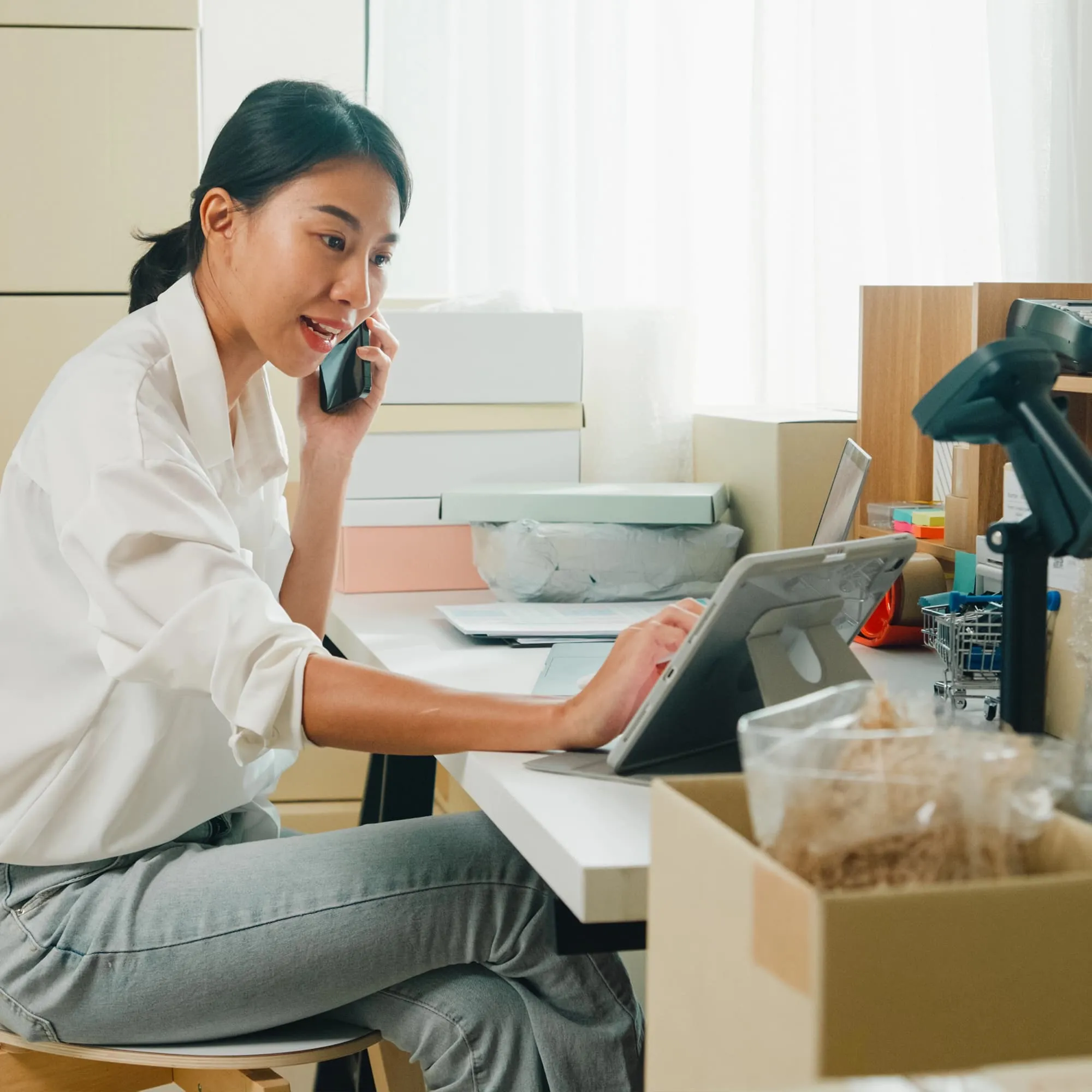 Woman talking on phone while working on a tablet at a desk with packaging boxes and supplies.