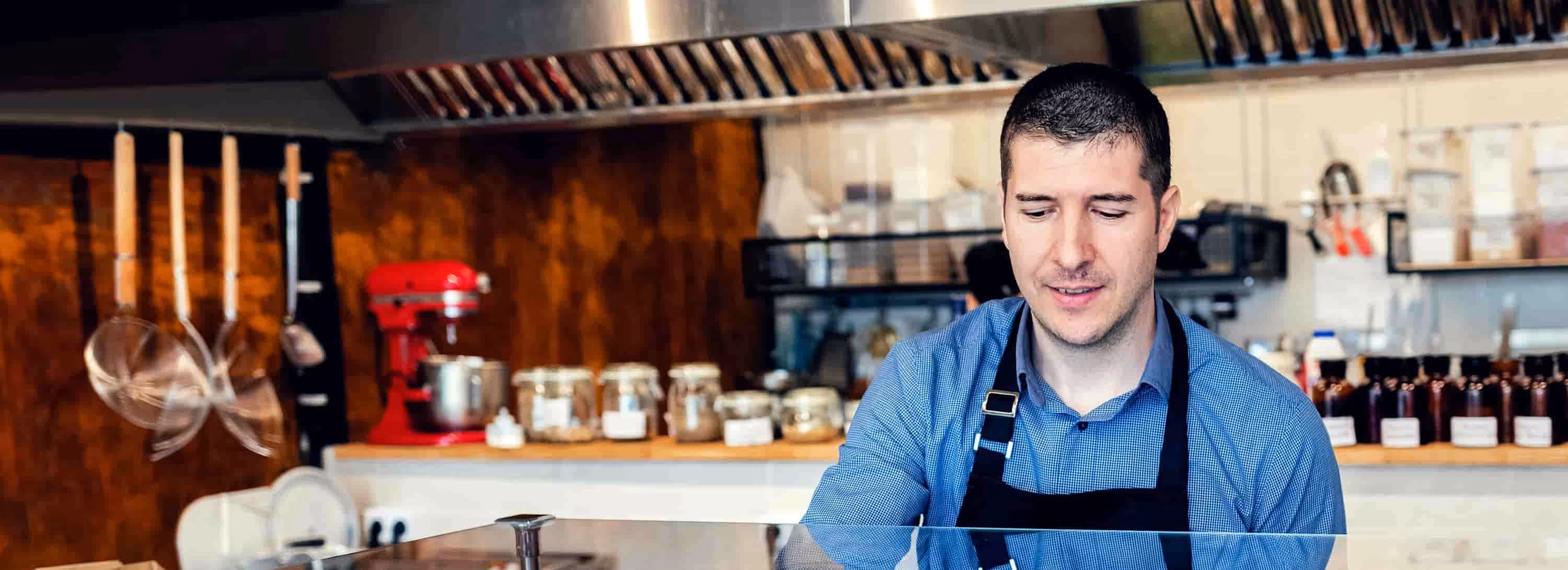 Man in apron working behind a glass counter in a kitchen with utensils and jars in background.