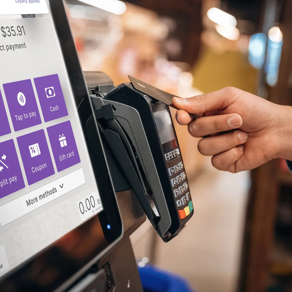 Hand holding a credit card tapping on a payment terminal next to a screen showing payment options.
