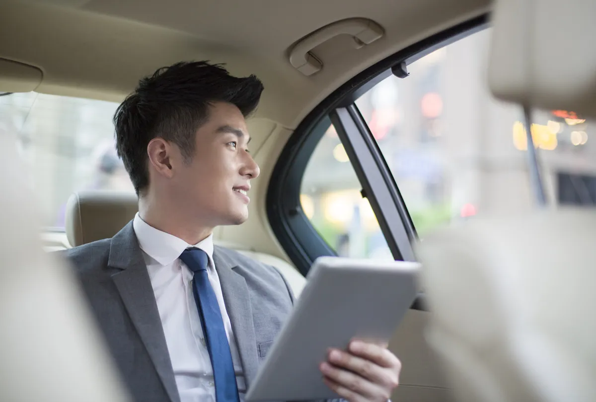 スーツを着た笑顔のビジネスマンがタブレットを持って車の後部座席に座って窓の外を見ている。