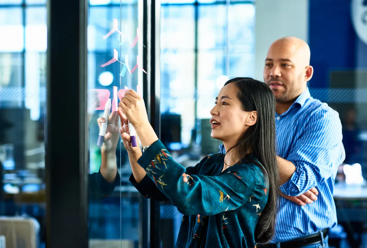 Woman writing on glass board with sticky notes while man watches in a modern office.