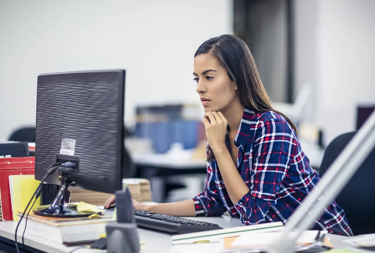 チェック柄のシャツを着た女性は、散らかったオフィスデスクでデスクトップコンピュータに集中していました。