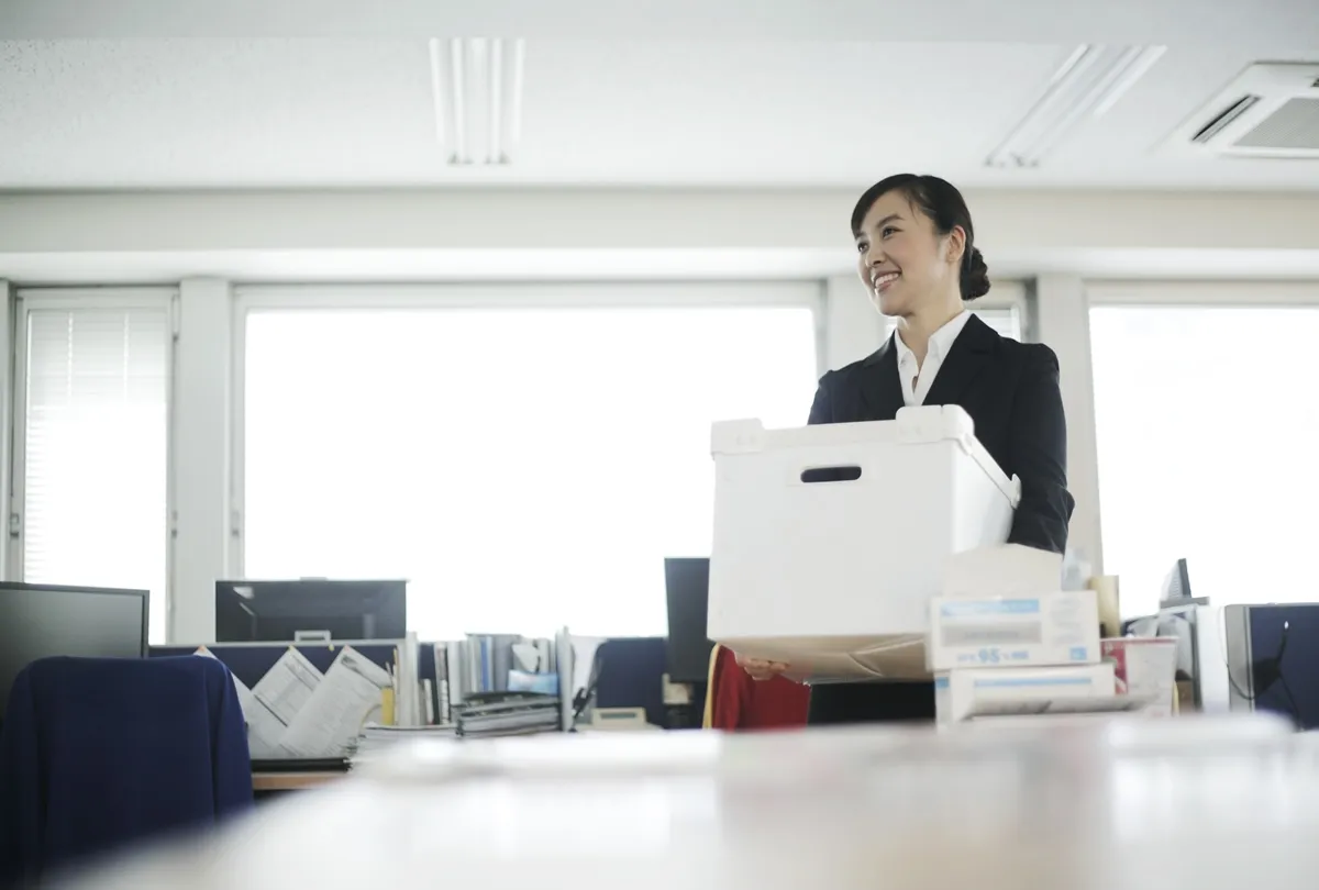 机とコンピューターのあるオフィス環境で白い箱を持ったビジネス服を着た笑顔の女性。