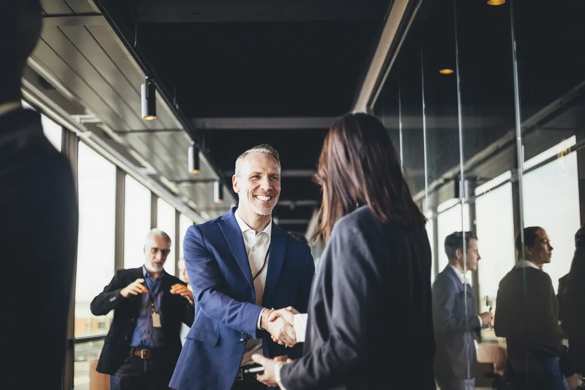 Man in a blue suit smiling and shaking hands with a woman in a business setting with others in background.