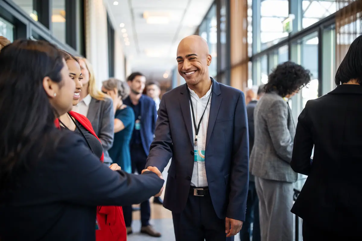 Smiling man in suit shaking hands with a woman in a busy office hallway during a professional event.