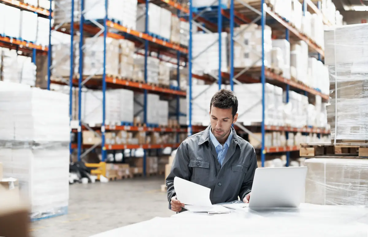 Man reviewing paperwork at a warehouse desk with laptop and stacked pallets in the background.