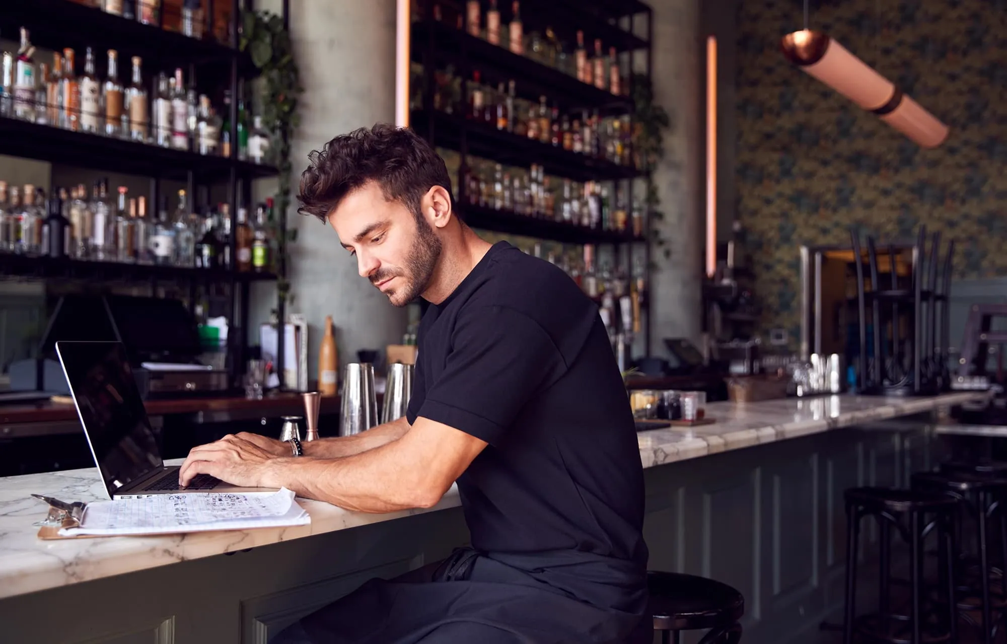 Man in black shirt sitting at bar counter working on a laptop with bottles on shelves behind him.