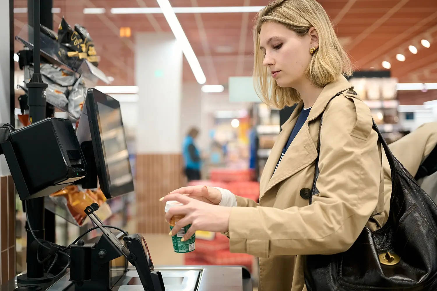 Woman with a beige coat and black bag using a self-checkout scanner in a grocery store.