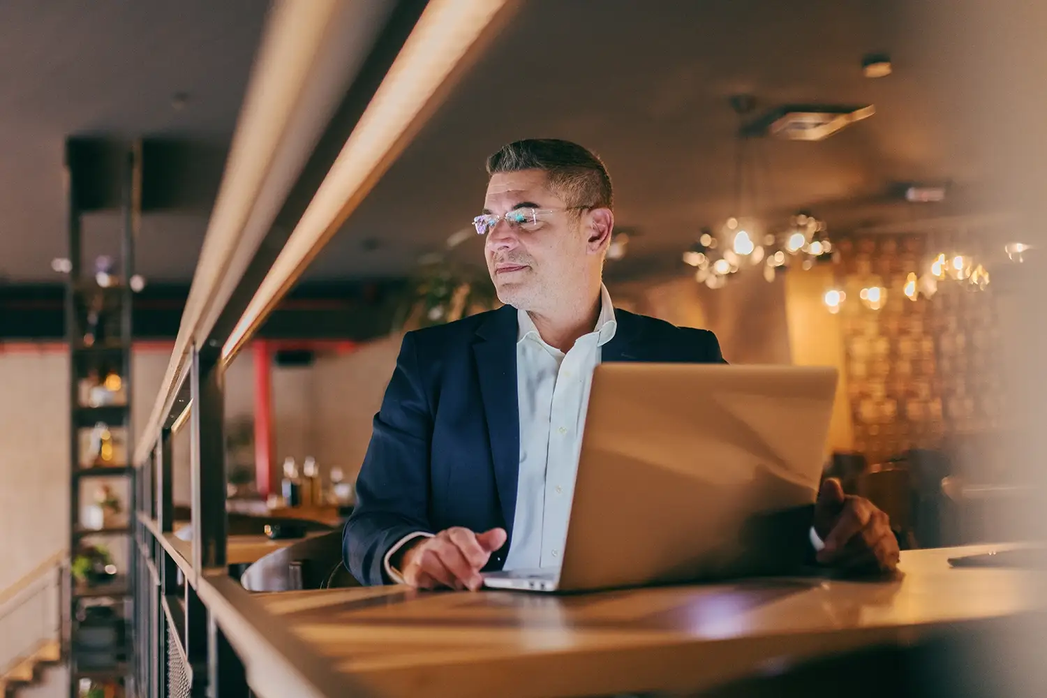 Man in glasses and suit looking sideways while using a laptop in a softly lit modern café.