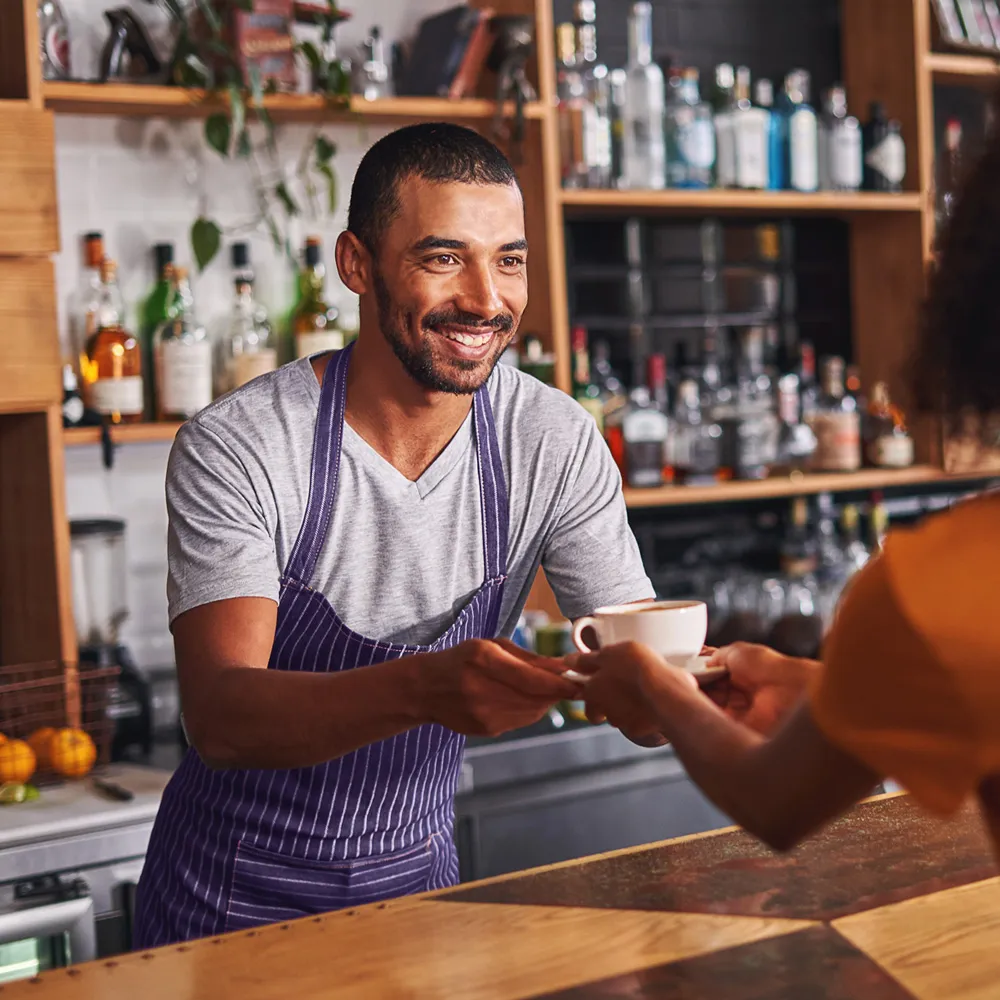 ストライプのエプロンを着た笑顔のバリスタが、カウンター越しに顧客にコーヒーを渡します。
