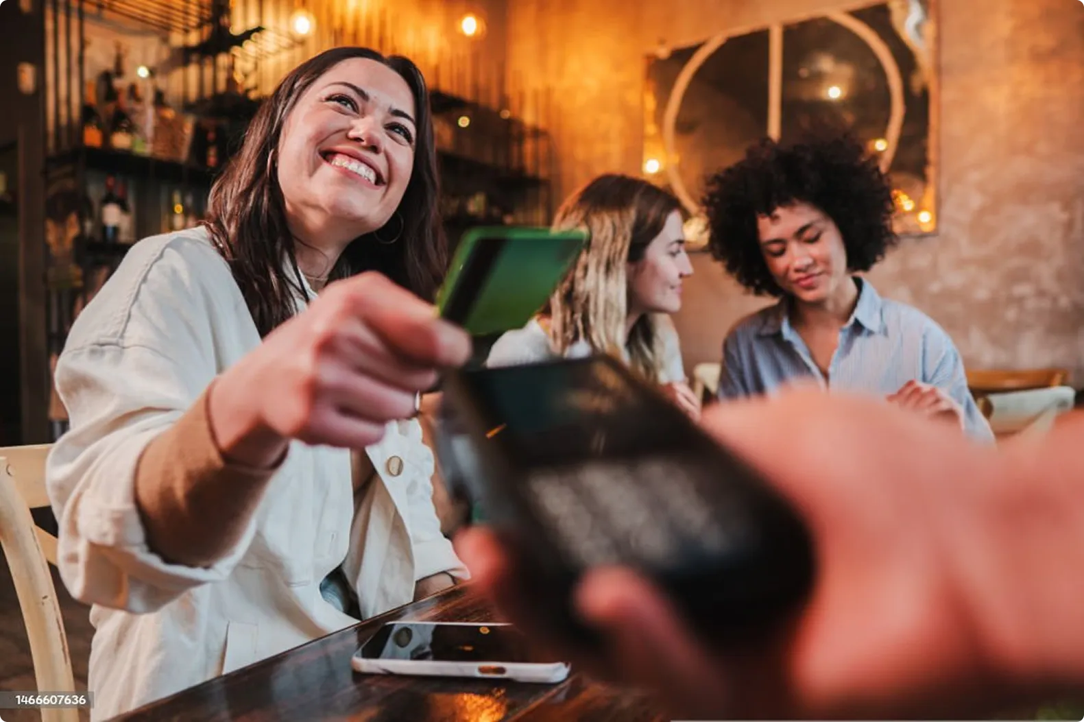Smiling woman paying for dinner with credit card at a table with friends in a cozy restaurant.