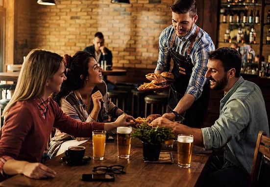 Waiter serving burgers to three friends sitting around a wooden table with drinks in a cozy restaurant.