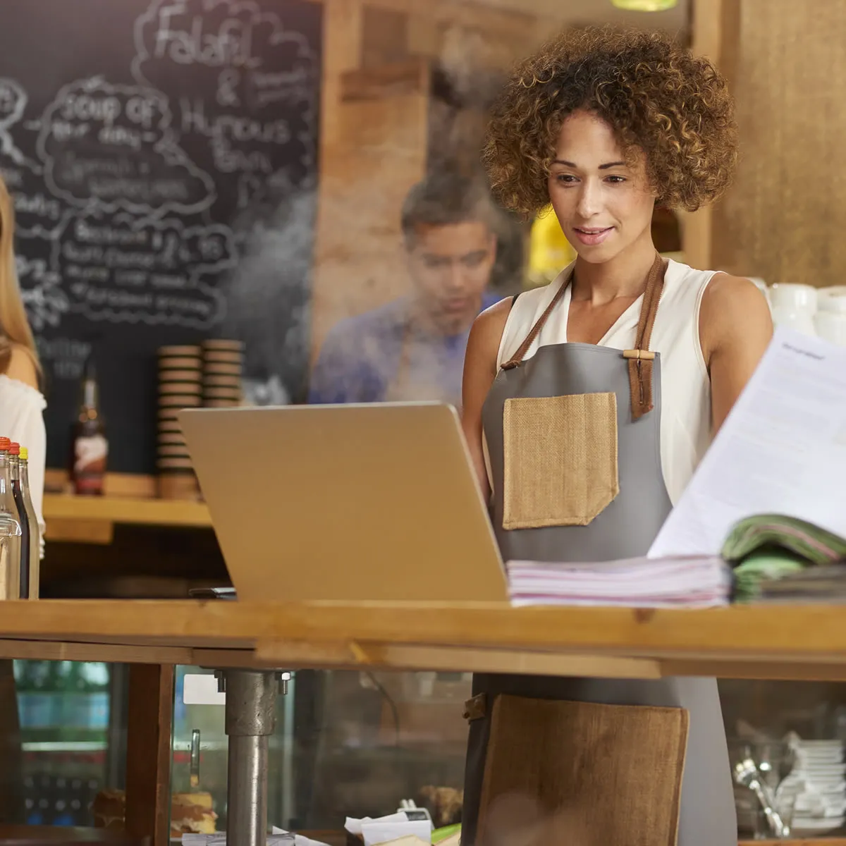 Female restaurant manager wearing apron reviewing reports and using laptop behind wooden counter.
