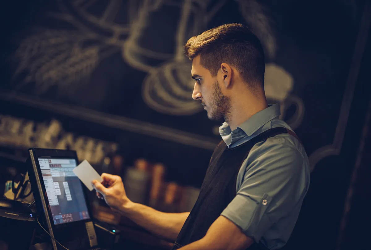 Man in blue shirt and black vest holding a card while using a touchscreen register in a café.