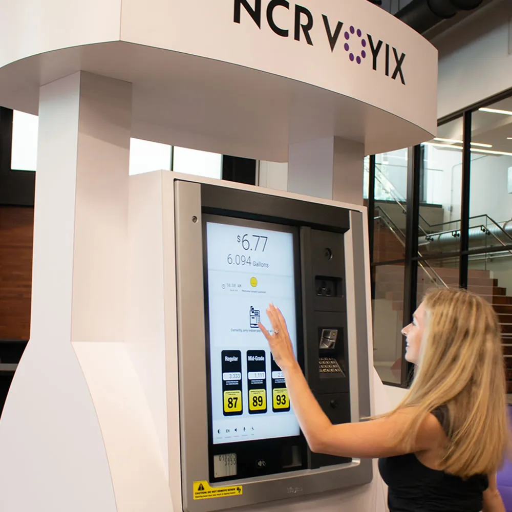 Woman using a large NCR Voyix touchscreen kiosk displaying fuel prices and gallons.
