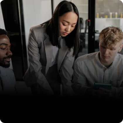 A woman in a gray blazer leans over a table discussing with two men, one holding a tablet, in a modern office setting.