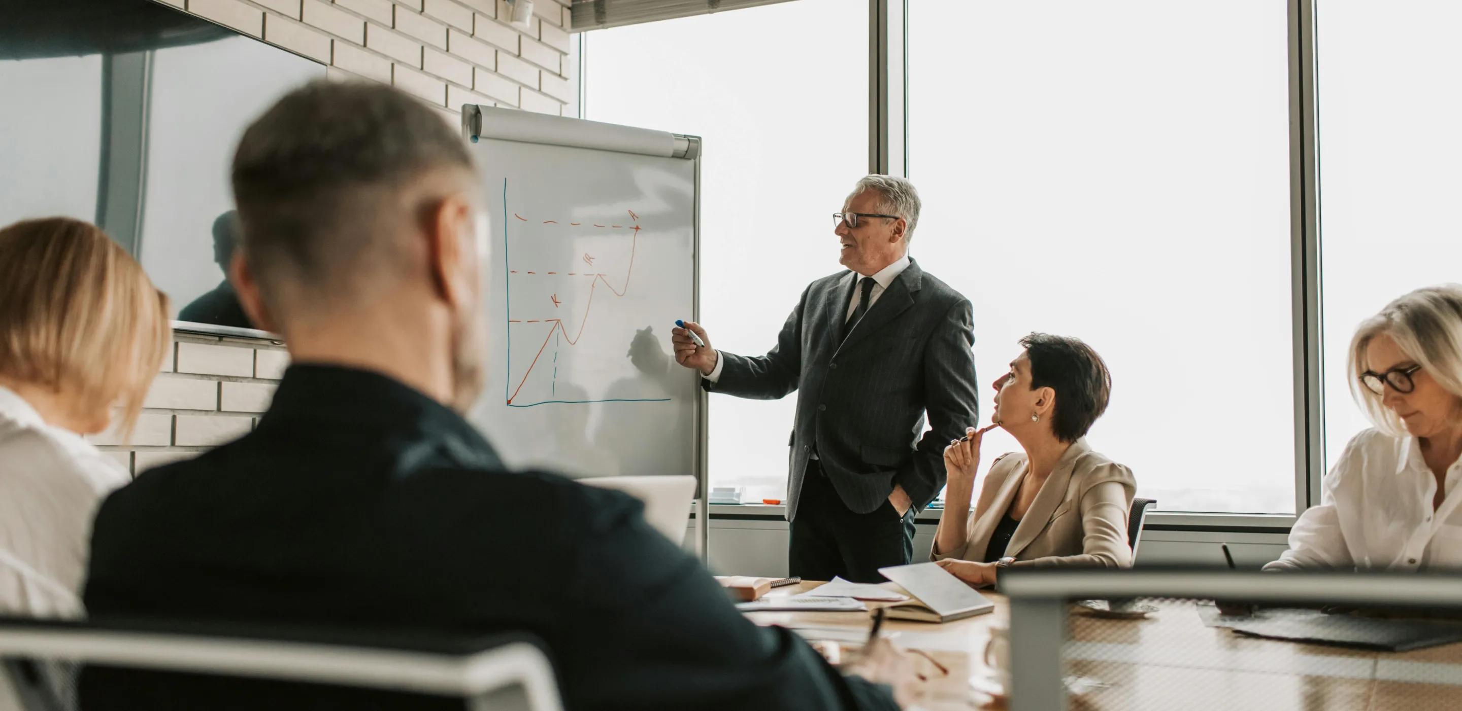 Businessman presenting a graph on a whiteboard to colleagues seated around a conference table in a modern office.