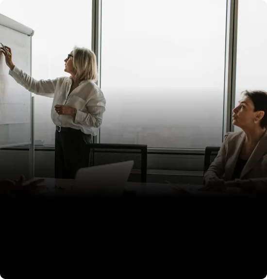 Two businesswomen in a meeting room, one writing on a flipchart and the other attentively watching.