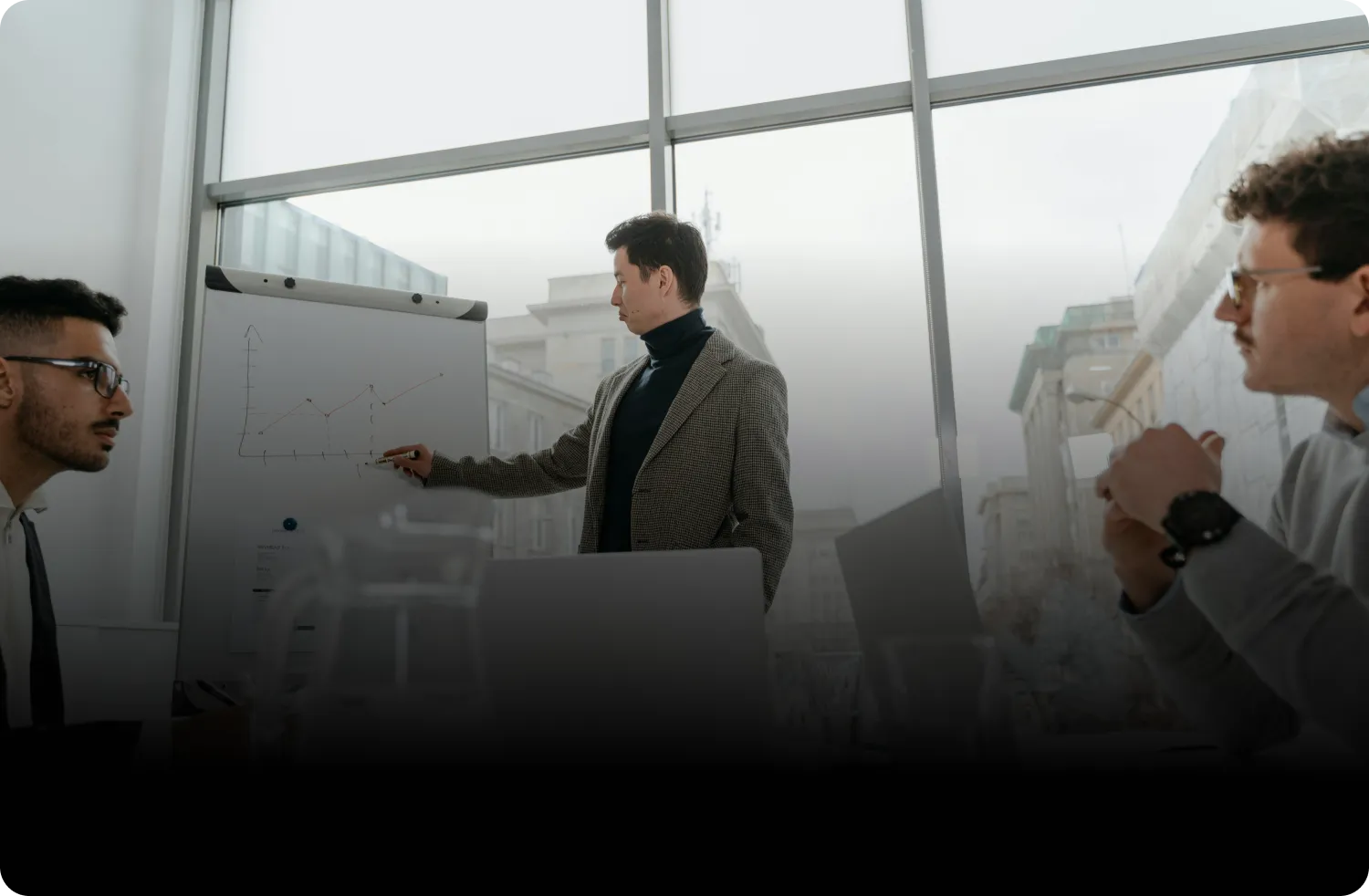 Man in a blazer presenting a line graph on a flip chart to two seated men with laptops in a bright office with large windows.