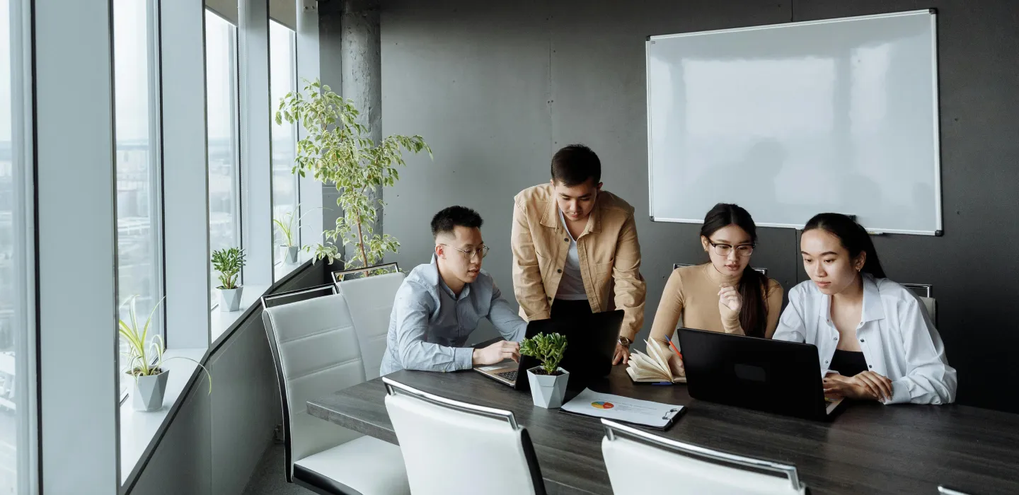 Four young professionals collaborating around a conference table with laptops and documents in a modern office with large windows and a whiteboard.