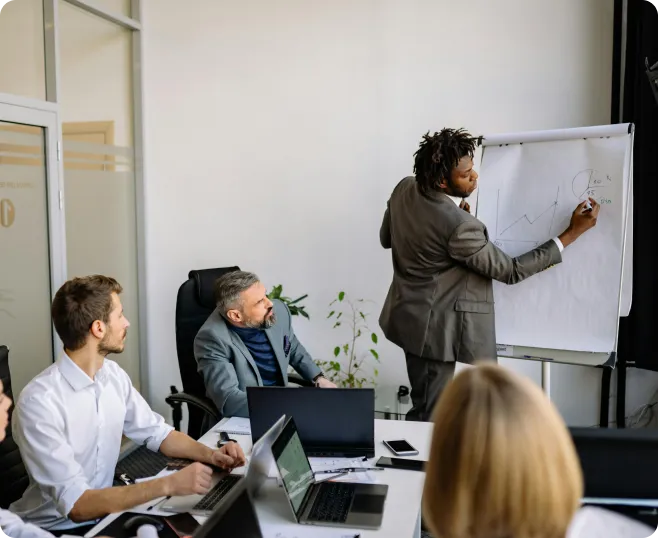 Businessman presenting a graph on a flip chart to colleagues seated at a conference table with laptops in a modern office.