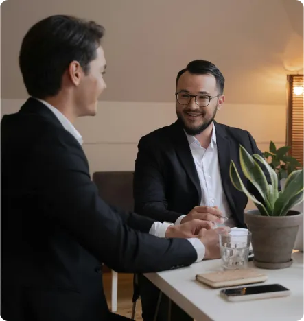 Two men in suits sitting at a table having a friendly business conversation with a potted plant and glasses of water in front of them.