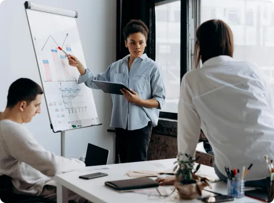 Woman presenting data charts on a flip chart to two colleagues in a modern office.