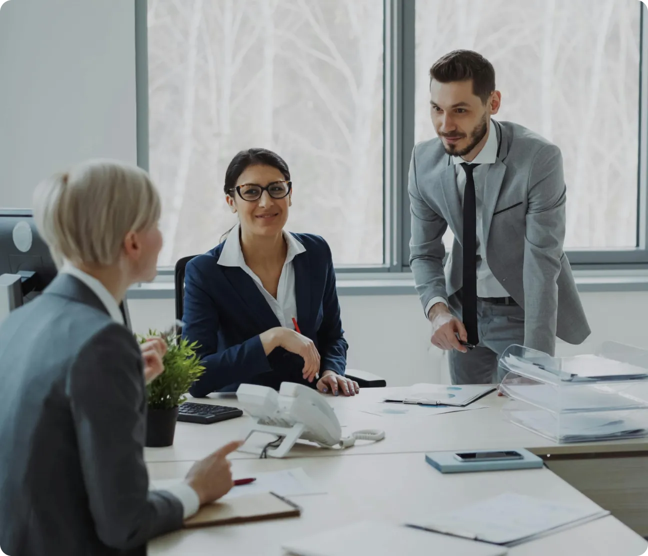 Three business professionals, two women and one man, engaged in a conversation around an office desk with documents and a telephone.