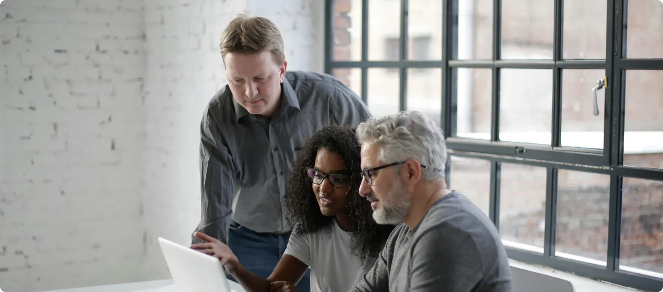 Three colleagues collaborating and looking at a laptop screen in a bright office with large windows.