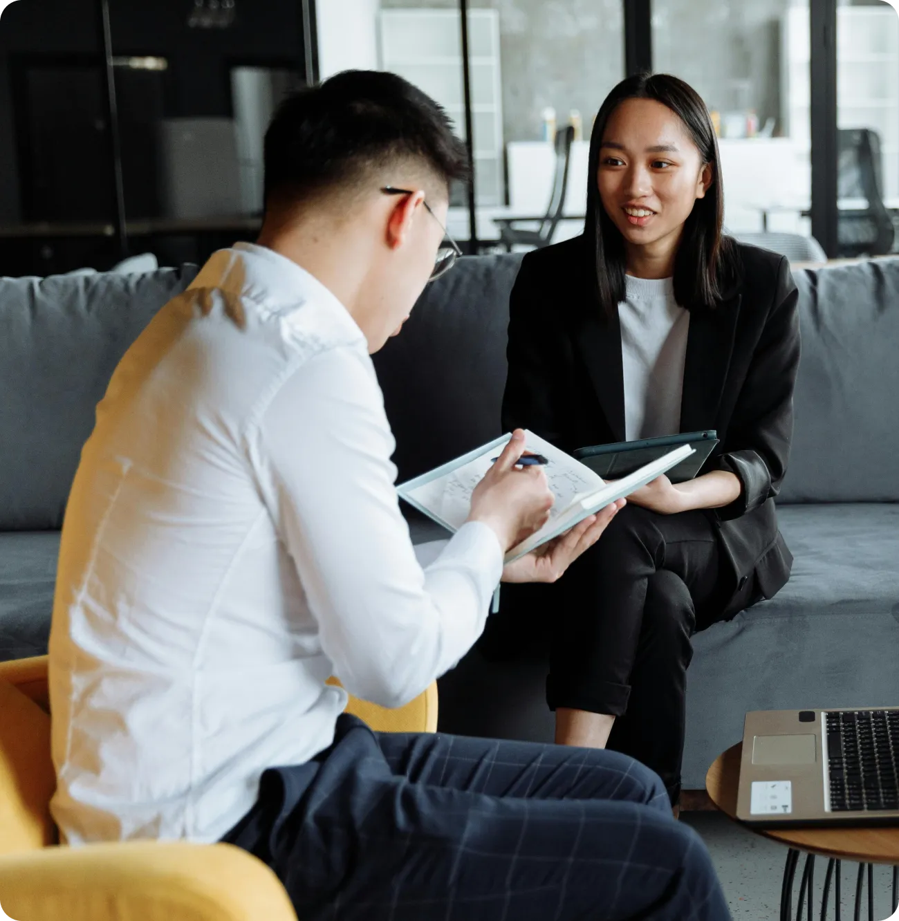 A man in a white shirt writing in a notebook while a woman in a black blazer smiles and listens during a meeting in a modern office.