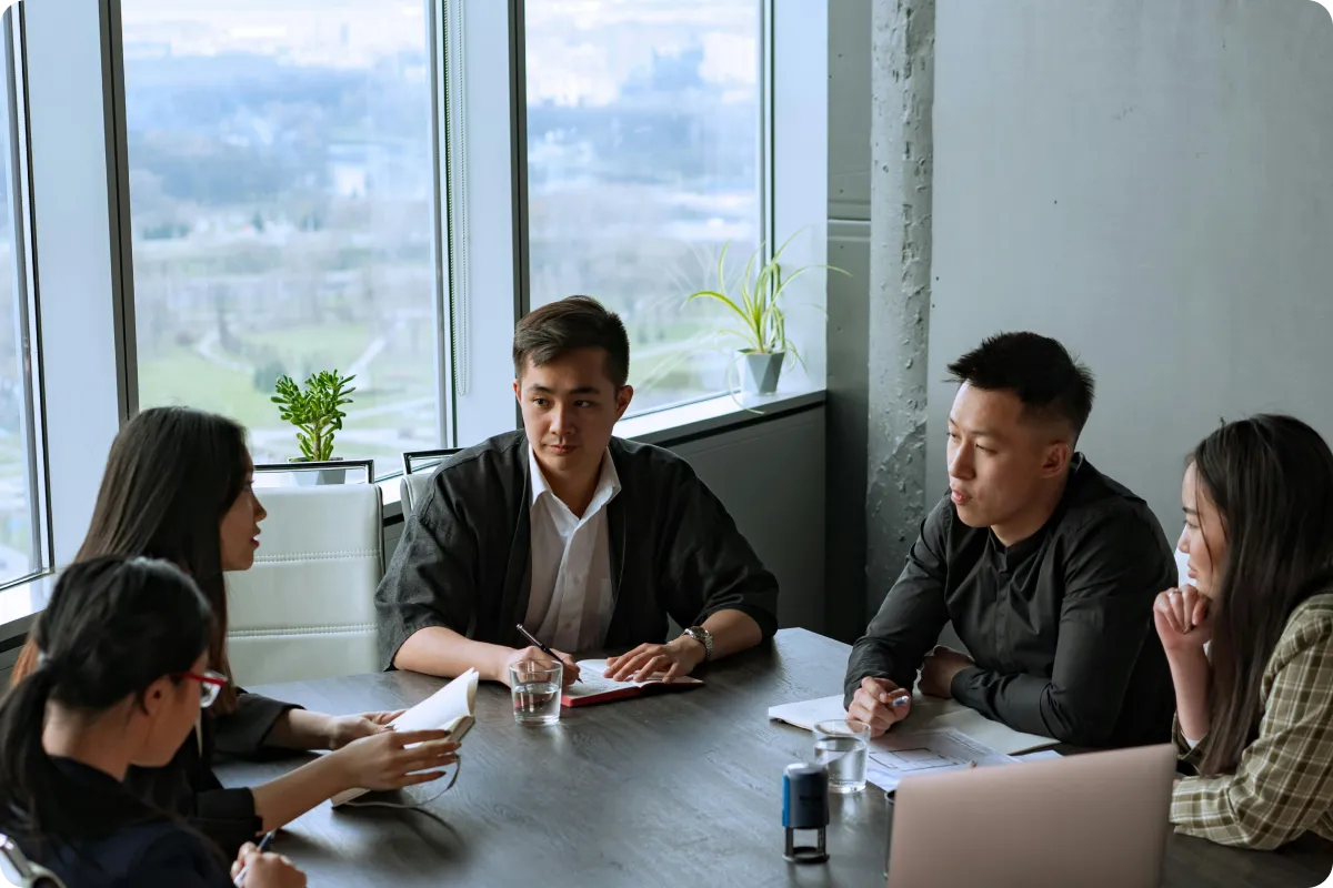 Five people having a meeting at a table with notebooks and glasses of water near large windows.
