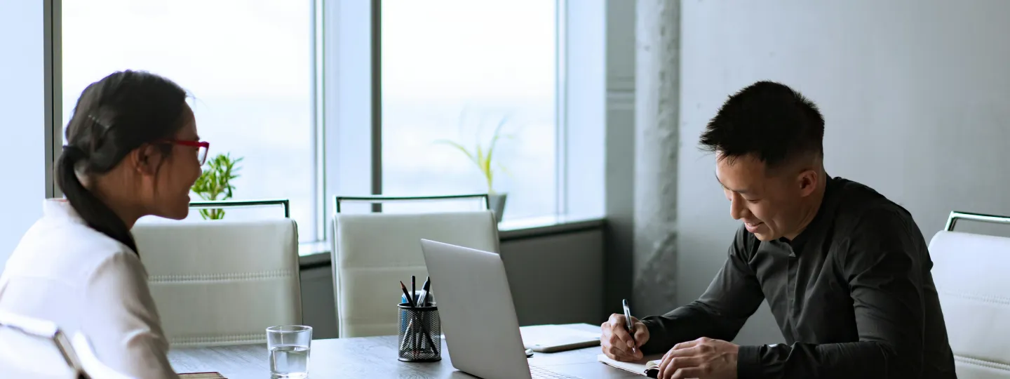 Two colleagues having a meeting in an office with large windows, one smiling woman and one man writing notes at a desk with a laptop.