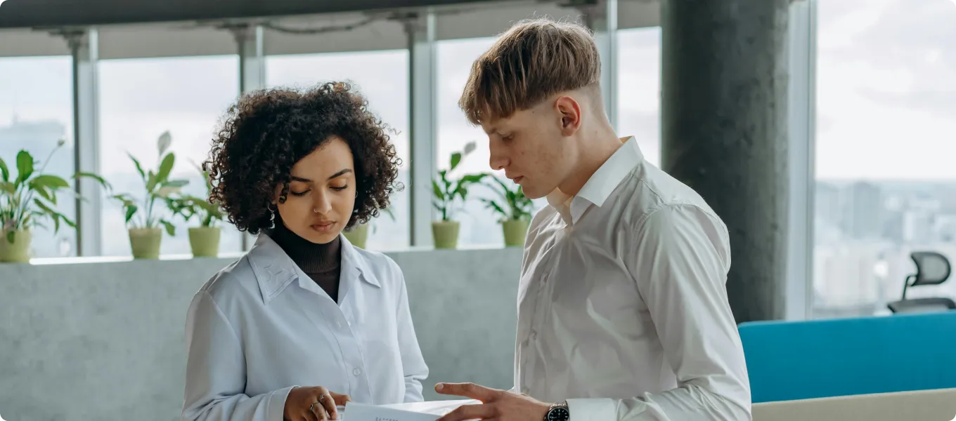 Two young professionals discussing documents in a modern office with large windows and potted plants.