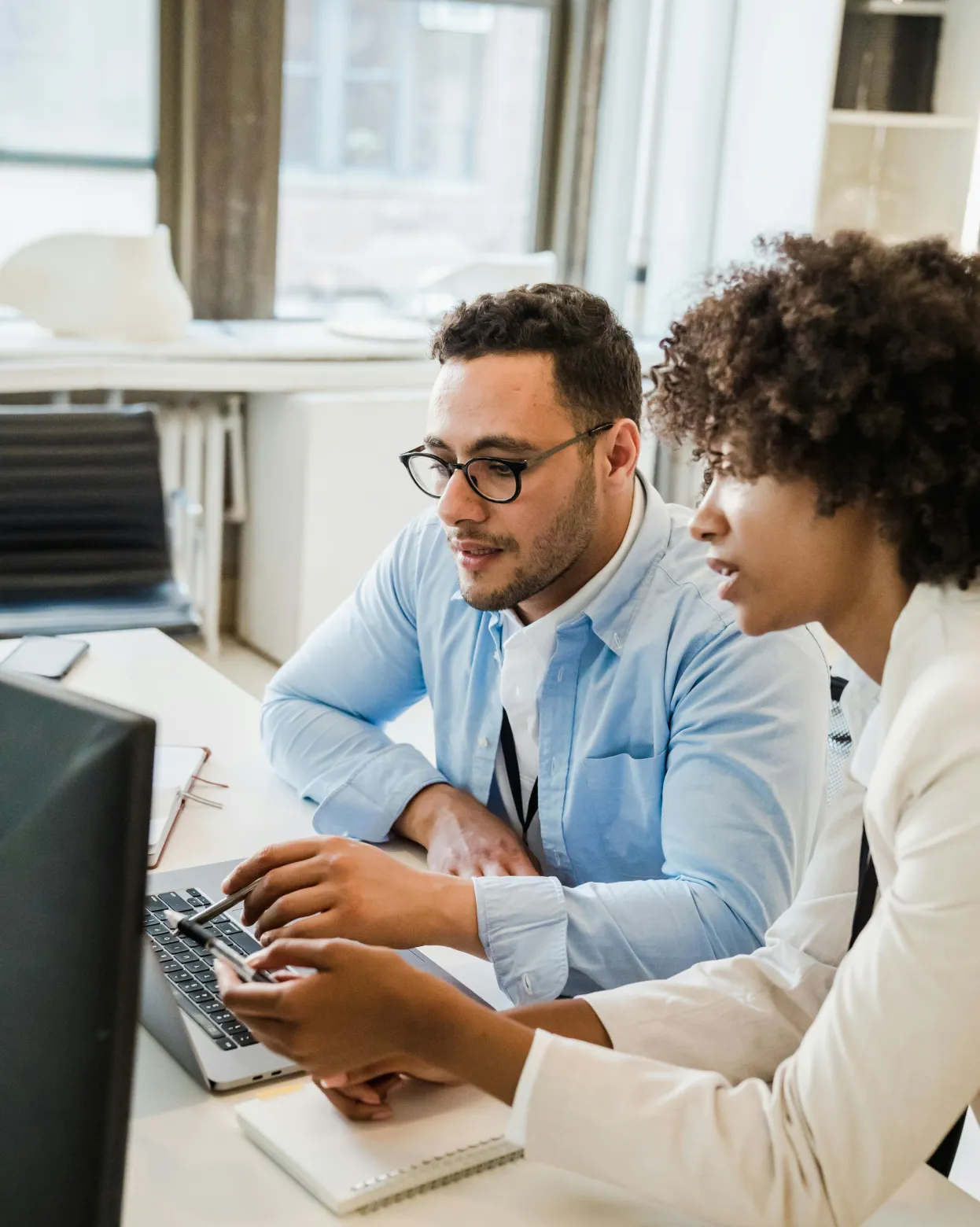 Two professionals, a man in glasses and a woman with curly hair, collaborating while looking at a laptop screen in a bright office.