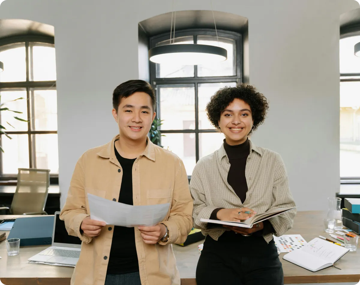 Two young professionals smiling in an office, one holding a paper and the other a notebook, standing in front of a desk with office supplies.