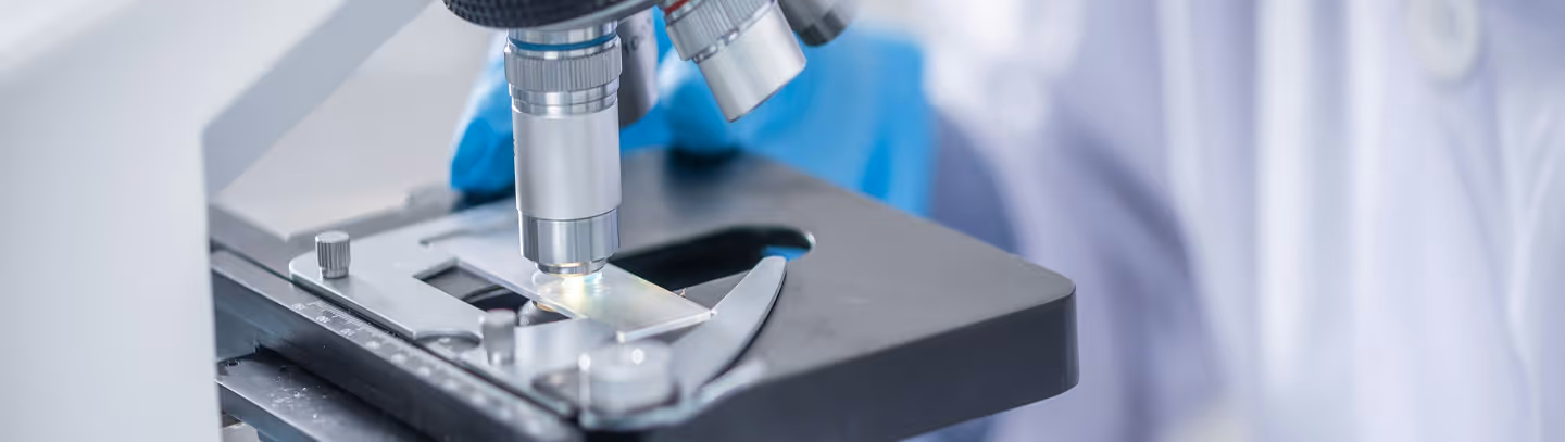 Gros plan sur un microscope avec une lame en verre examinée, une personne portant des gants bleus en arrière-plan.