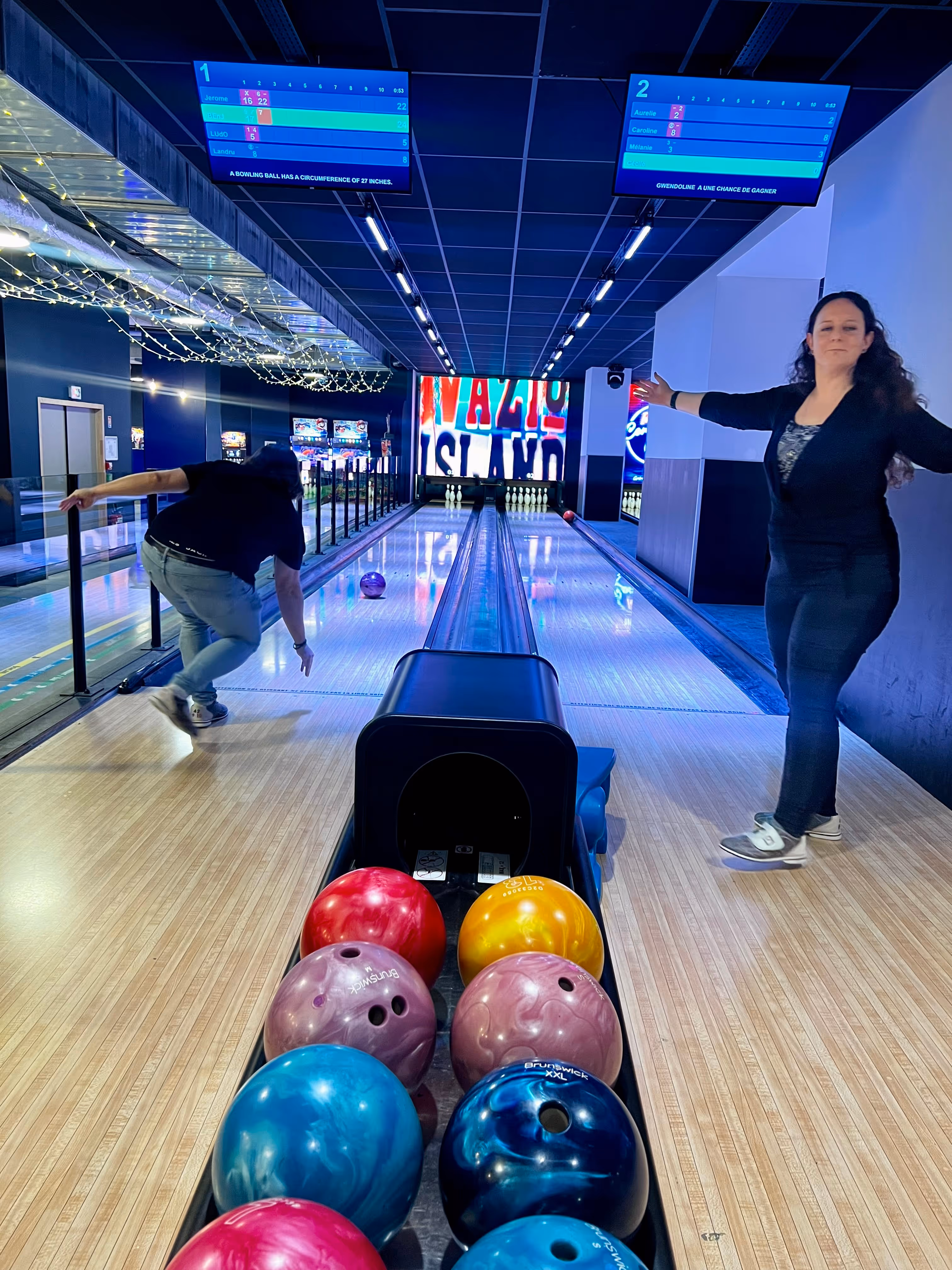 Deux personnes jouent au bowling dans une piste intérieure avec plusieurs boules de bowling colorées visibles au premier plan.