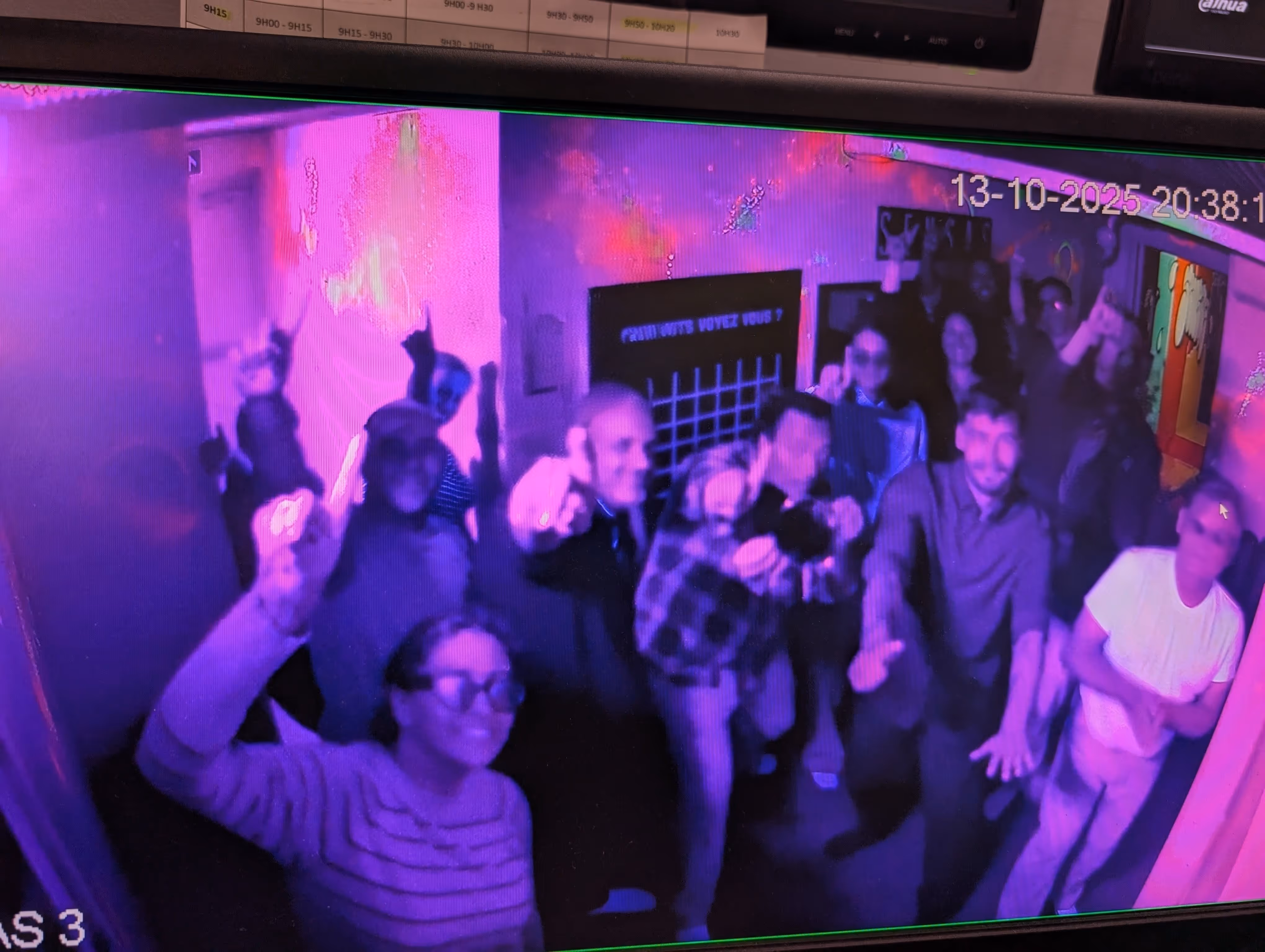 Groupe de personnes dans une pièce avec lumière violette faisant des gestes de célébration vers une caméra de surveillance datée du 13 octobre 2025.